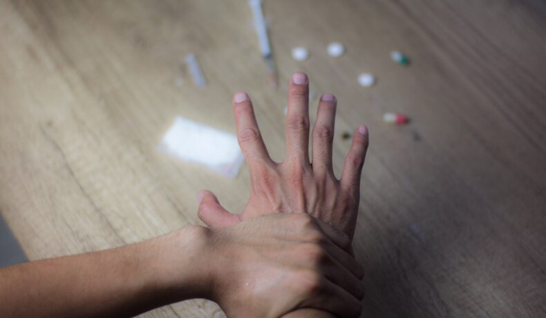 Symboliczna scena walki z uzależnieniem od dopalaczy – jedna ręka powstrzymuje drugą przed sięgnięciem po narkotyki, strzykawki i tabletki leżące na stole